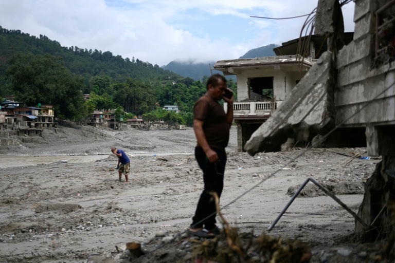 Deux hommes au téléphone au milieu d'habitations recouvertes de boue et de débris après la crue destructrice qui a fait plus de 600 morts à la frontière entre le Népal et la Chine, le 29 août 2026 à Devighat, dans le district népalais  de Nuwakot 