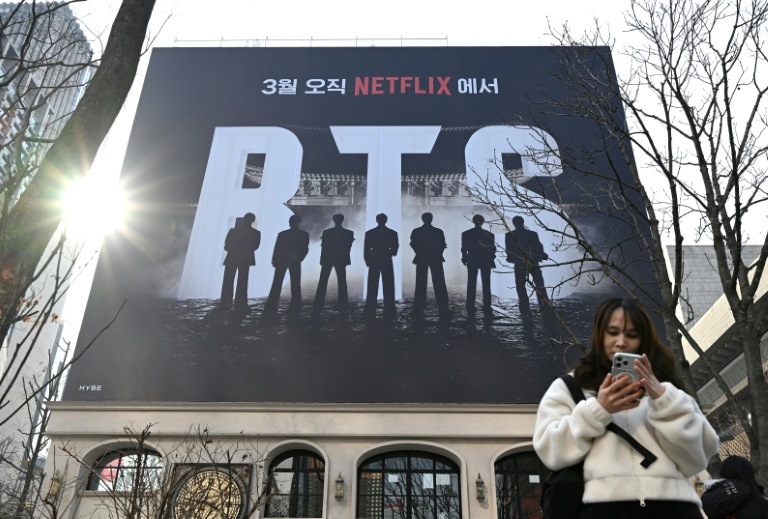 Une femme consulte son smartphone devant un panneau d'affichage annonçant le concert de retour du groupe de K-pop BTS, le 17 mars 2026 à Séoul
