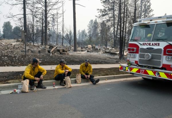 Trois membres du service d'incendie de Spokane en pause sur le bord du trottoir devant des maisons d'habitation incendiées, dans le quartier de Balboa à Spokane (Washington), le 2 août 2026