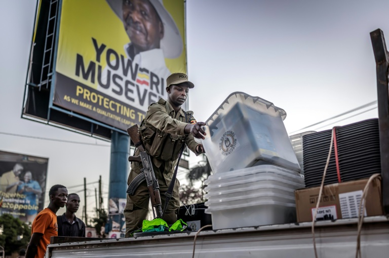 Un policier décharge d'un camion des urnes destinés à un bureau de vote devant une affiche électorale du président sortant Yoweri Museveni, candidat du Mouvement de résistance nationale (NRM) à la présidentielle, le 15 janvier 2026 à Kampala, en Ouganda